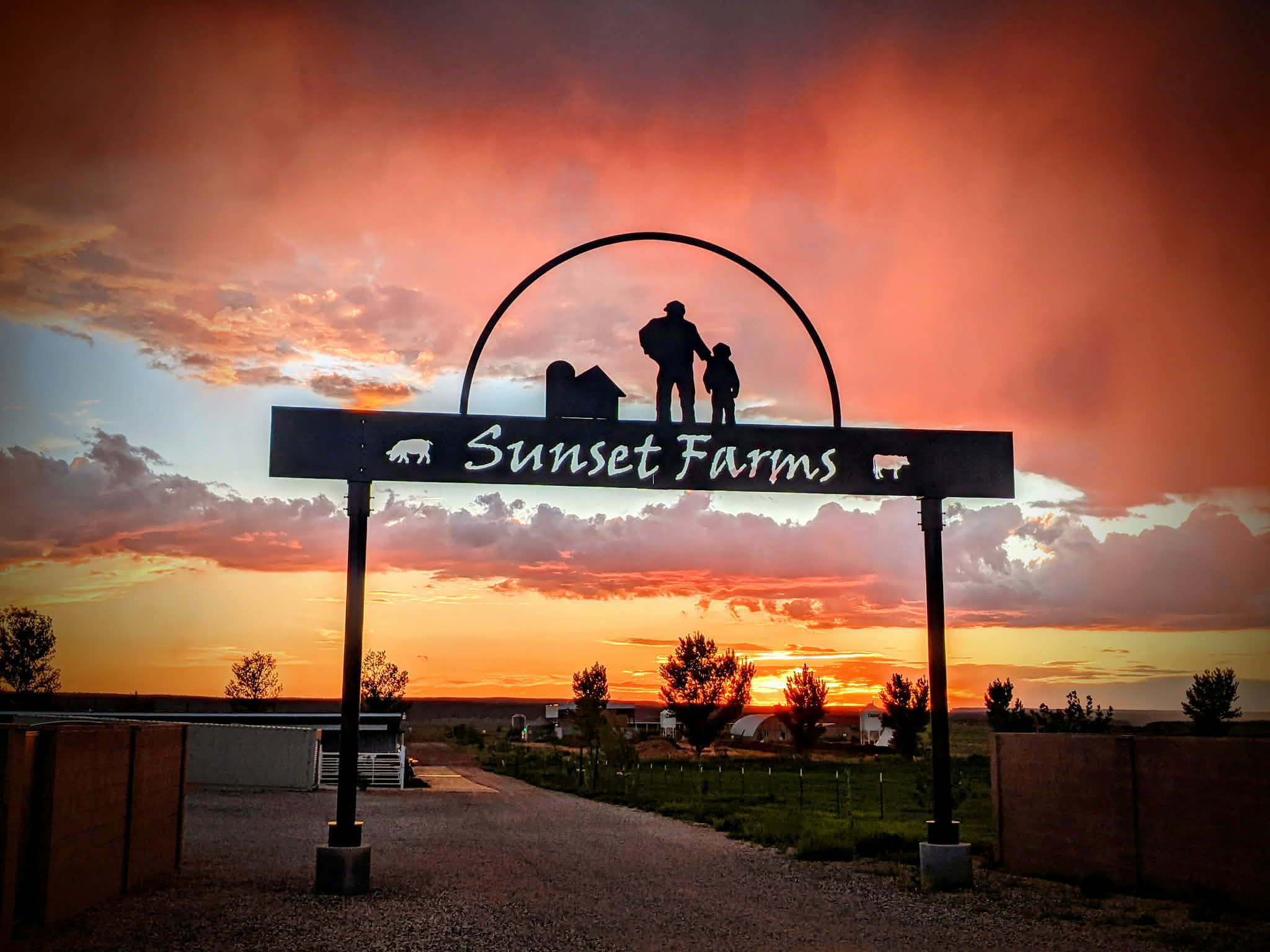 The Sunset Farms entrance gate at sunset, family silhouette and barn under fiery sky over the home pasture